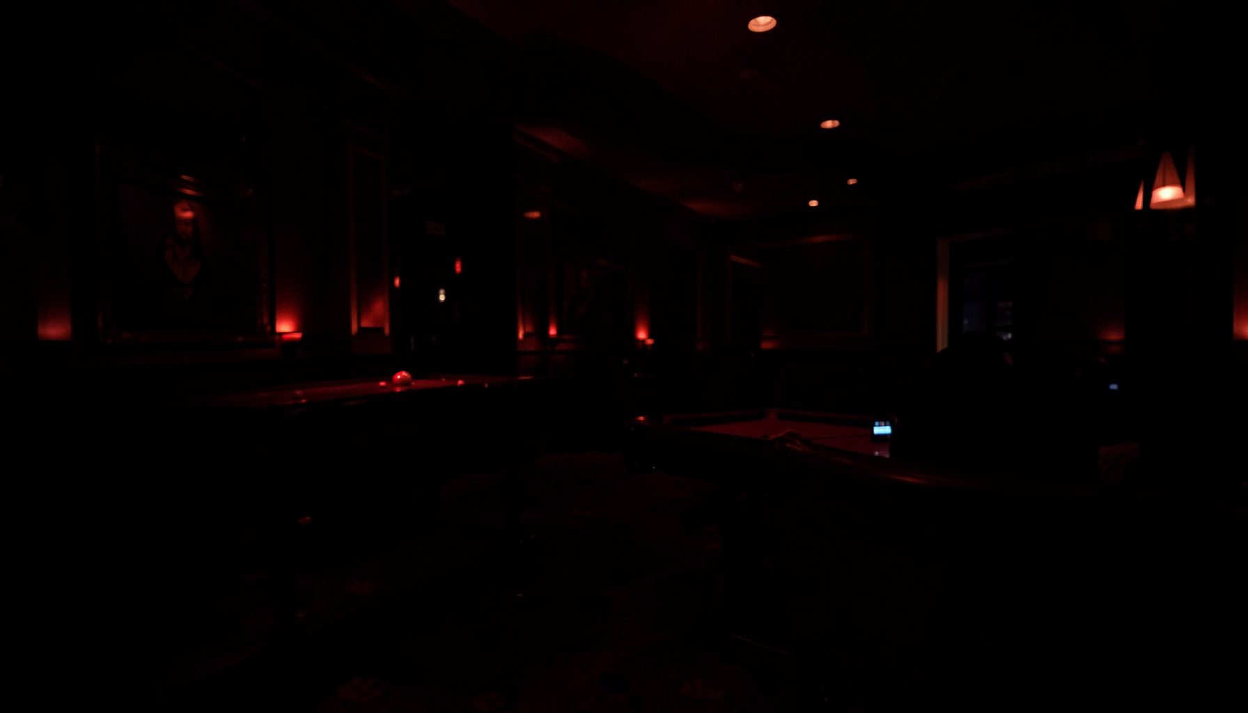 Dark, low-key photograph of a dimly lit casino interior, deep red and black tones with soft ambient light reflecting off polished surfaces, moody and quiet atmosphere.