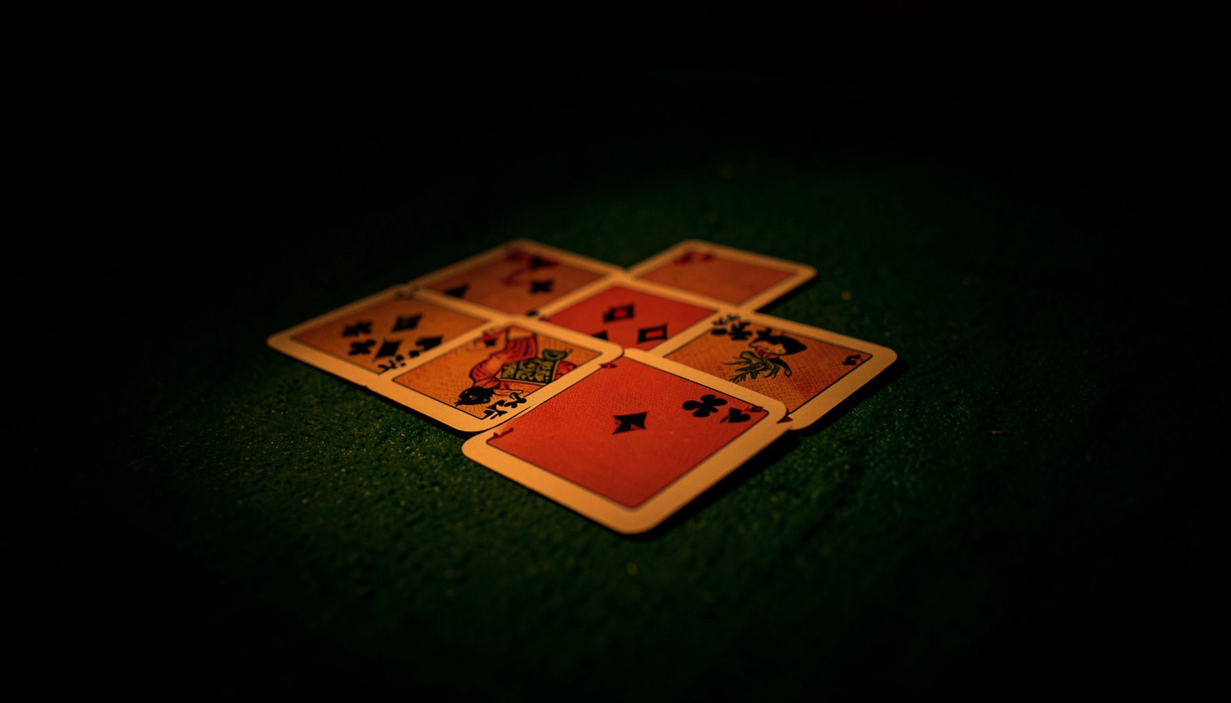 A spread of playing cards on a green felt surface under moody, low amber lighting with deep shadows around the edges