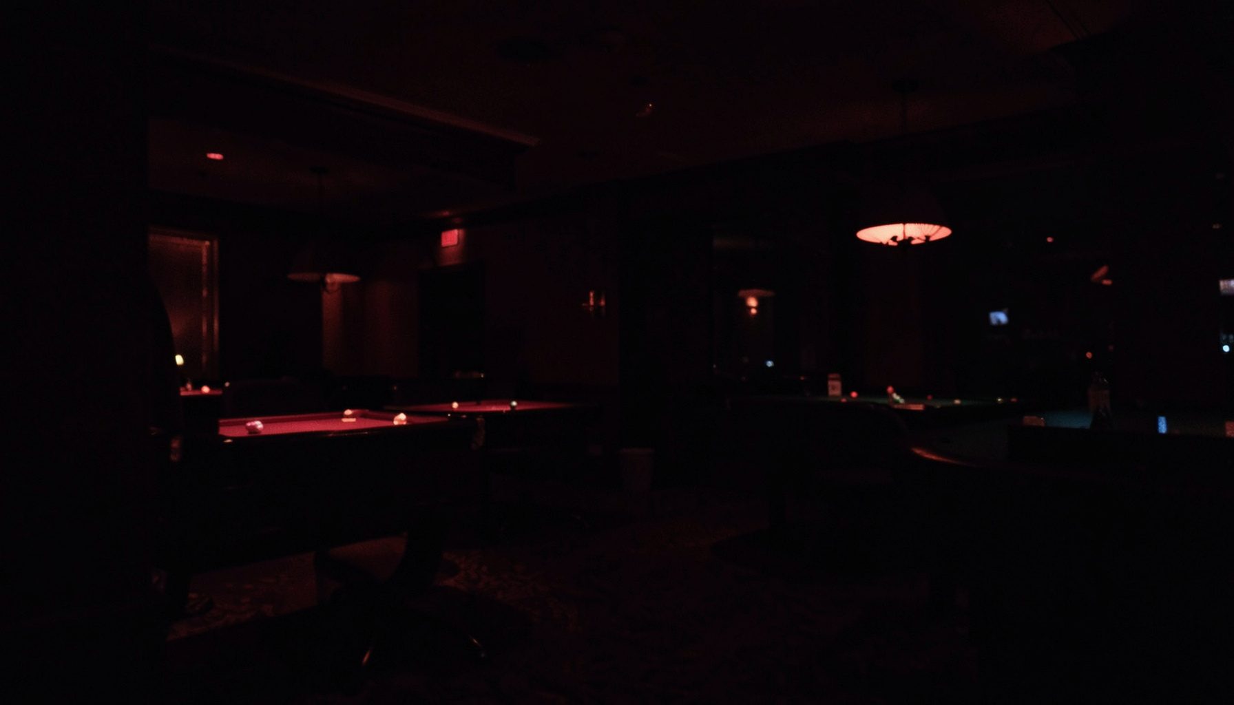 Dark moody photograph of a dimly lit casino interior with deep maroon and shadow tones, faint glow from card tables in the background